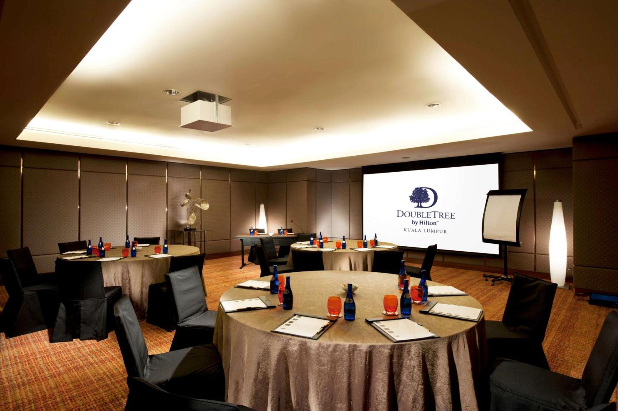 Doubletree By Hilton Kuala Lumpur Hotel Kültér fotó The photo shows a conference room set up for a meeting or event. There are several round tables with beige tablecloths, each adorned with blue and red decorations, likely drinks and supplies. Black chairs are placed around the tables. On one wall, th